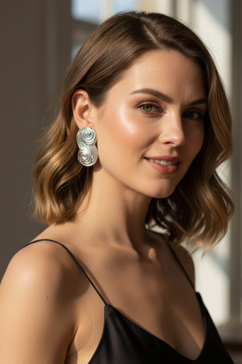 Side view of a model with wavy hair wearing the Seraphina silver sculptural earrings. The high-polish finish reflects natural light, highlighting the bold S-shape swirl and modern jewelry aesthetic.
