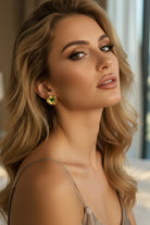 Glowy cinematic portrait of a blonde woman with wavy hair wearing a liquid metal aesthetic gold statement stud with a dark green emerald stone. Styled with a satin slip dress for summer resort wear or an outdoor cocktail party look.