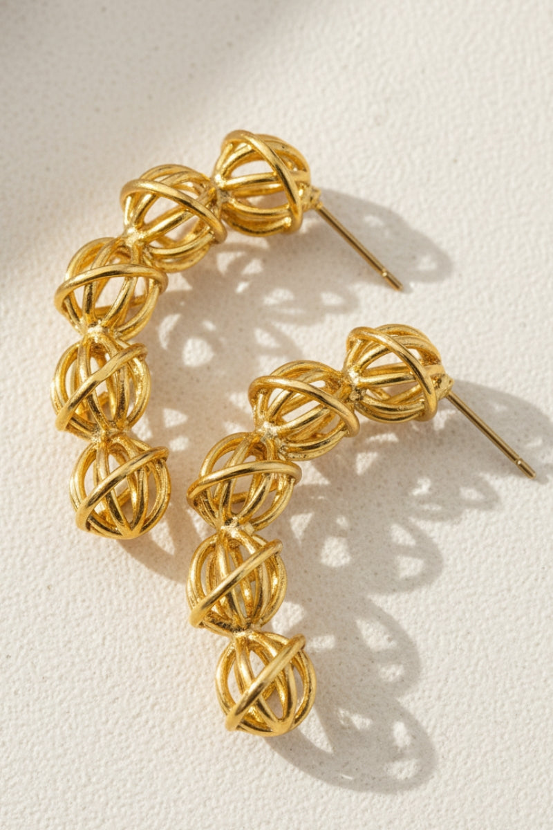 A warm-toned product flat lay showing the textured mesh metal spheres of the tarnish-resistant demi-fine gold hoops on a sand-colored surface, emphasizing artisanal craftsmanship and premium boutique quality.