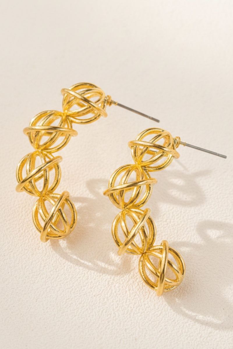 A pair of wire-woven gold curved drop earrings displayed on a soft textured cream surface under warm studio lighting, highlighting the intricate openwork cages and premium tarnish-free finish suitable for boutique designer collections.